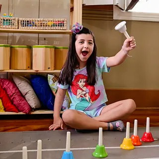 Menina sorri e ergue um sino sentada no chão da sala